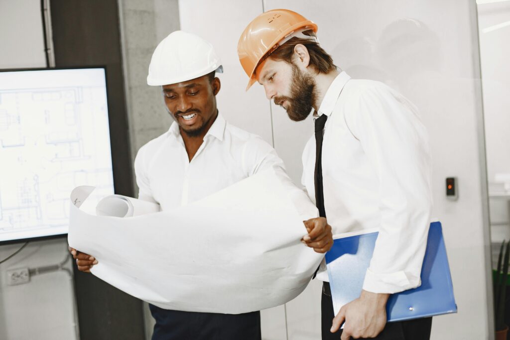 Two engineers in hard hats review construction blueprints in an office setting.